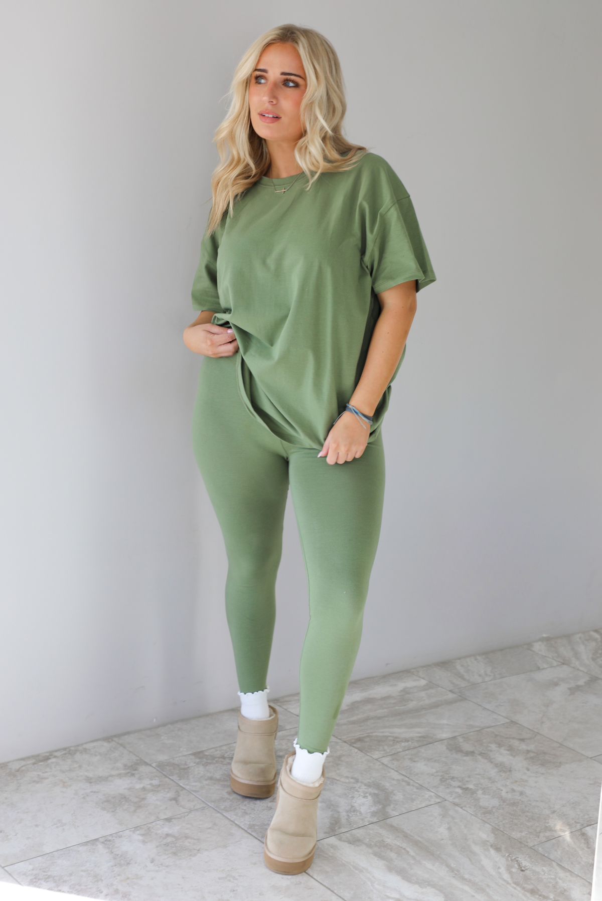 Woman wearing a green two piece set featuring an oversized t-shirt and fitted leggings. The set is paired with high white ankle socks and sand colored warm boots. The photo is taken indoors against a white wall with white marble flooring. 