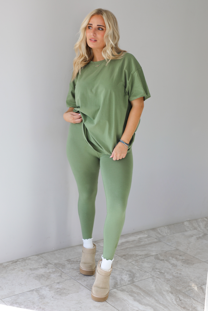 Woman wearing a green two piece set featuring an oversized t-shirt and fitted leggings. The set is paired with high white ankle socks and sand colored warm boots. The photo is taken indoors against a white wall with white marble flooring. 