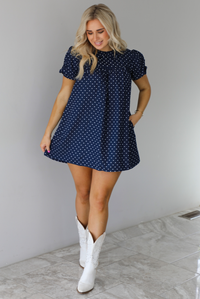 Woman wearing a navy baby-doll cut dress with a white polkadot print pattern throughout. The dress has puffed short sleeves and functional pockets on either side. The dress is paired with white cowboy boots. The photo is taken indoors against a white wall with white marble flooring. 