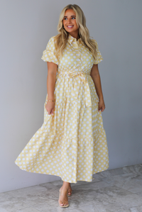 A woman wearing a yellow and white polka dot midi dress with puff sleeves and a tie waist and functional button closures along the front, standing indoors against a white wall with white marble flooring. 