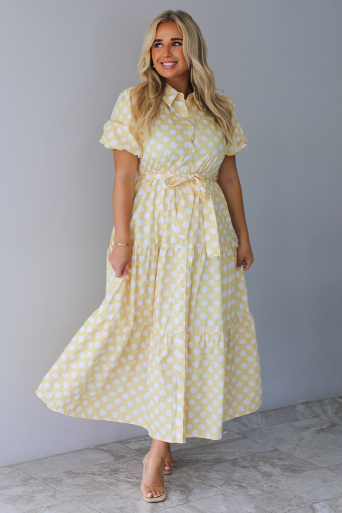 A woman wearing a yellow and white polka dot midi dress with puff sleeves and a tie waist and functional button closures along the front, standing indoors against a white wall with white marble flooring. 