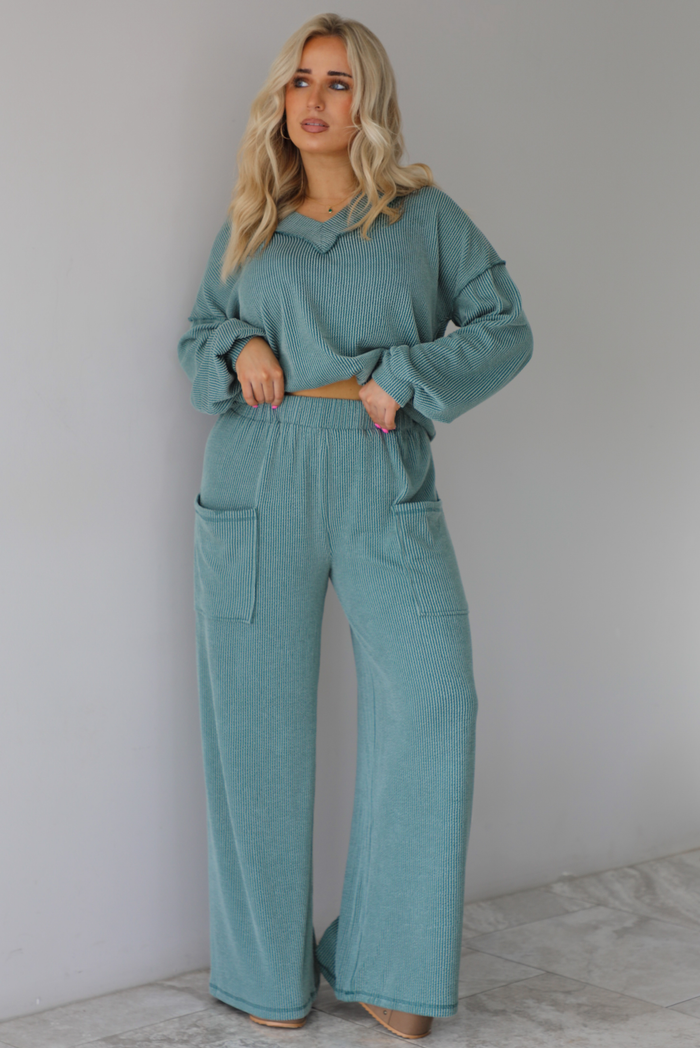 Woman wearing a teal two piece set that has a v-neck long sleeved oversized top and high waisted pants. The top is tucked into the woman's bra to give it a cropped look. The photo is taken indoors against a white wall with white marble flooring. 