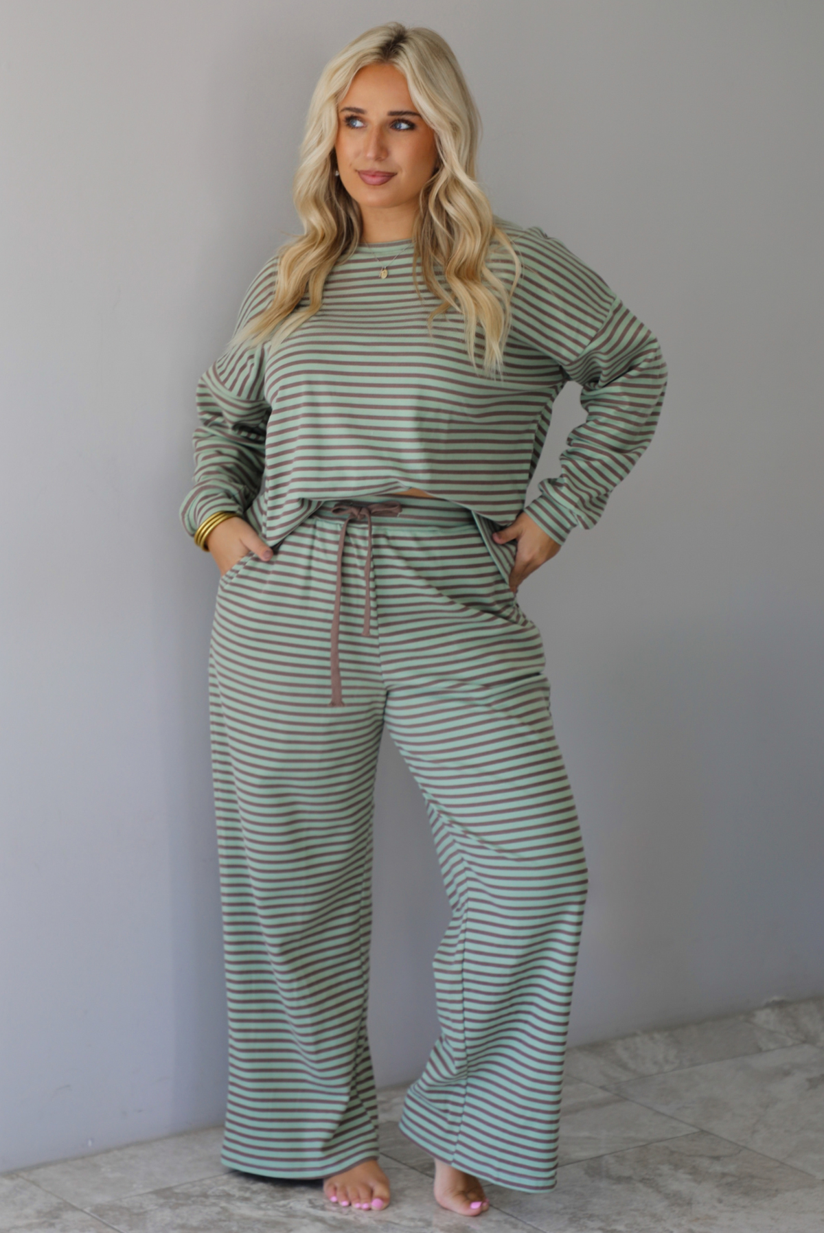 A woman wearing a pale green and brown striped two piece set featuring an oversized long sleeved top and high waisted, wide leg pants, standing indoors against a white wall with white marble flooring. 