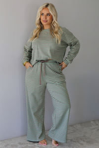 A woman wearing a pale green and brown striped two piece set featuring an oversized long sleeved top and high waisted, wide leg pants, standing indoors against a white wall with white marble flooring. 