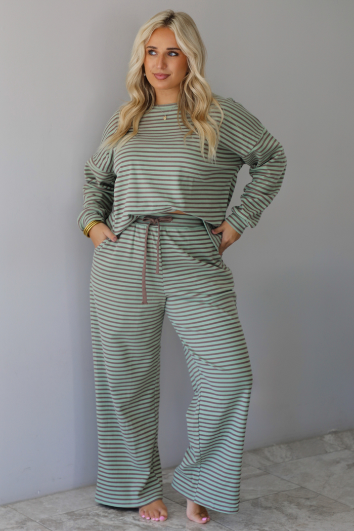 A woman wearing a pale green and brown striped two piece set featuring an oversized long sleeved top and high waisted, wide leg pants, standing indoors against a white wall with white marble flooring. 