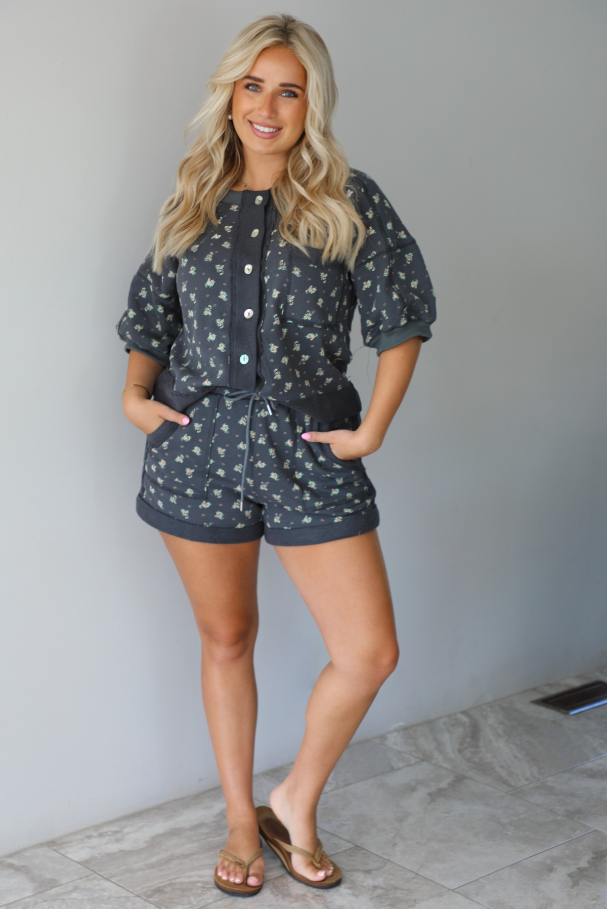 Woman wearing a charcoal with a multi-colored floral two piece set featuring an oversized top with puffed sleeves and shorts indoors against a white wall with white marble flooring. The top is tucked into the from of the shorts and the models hands are in the pockets of the shorts. 