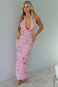 Woman wearing a formal pink maxi dress that has a plunging slouchy neckline and floral rhinestone details throughout. The photo is taken indoors against a white wall with white marble flooring. 
