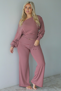 A woman wearing a maroon and white striped two piece matching set featuring a slouchy off the shoulder long sleeved top and mid-rise, folded over waistline wide leg pants, standing indoors against a white wall with white marble flooring. 