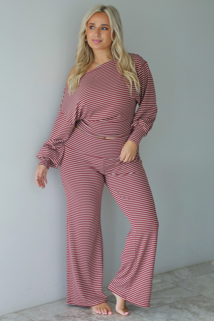 A woman wearing a maroon and white striped two piece matching set featuring a slouchy off the shoulder long sleeved top and mid-rise, folded over waistline wide leg pants, standing indoors against a white wall with white marble flooring. 