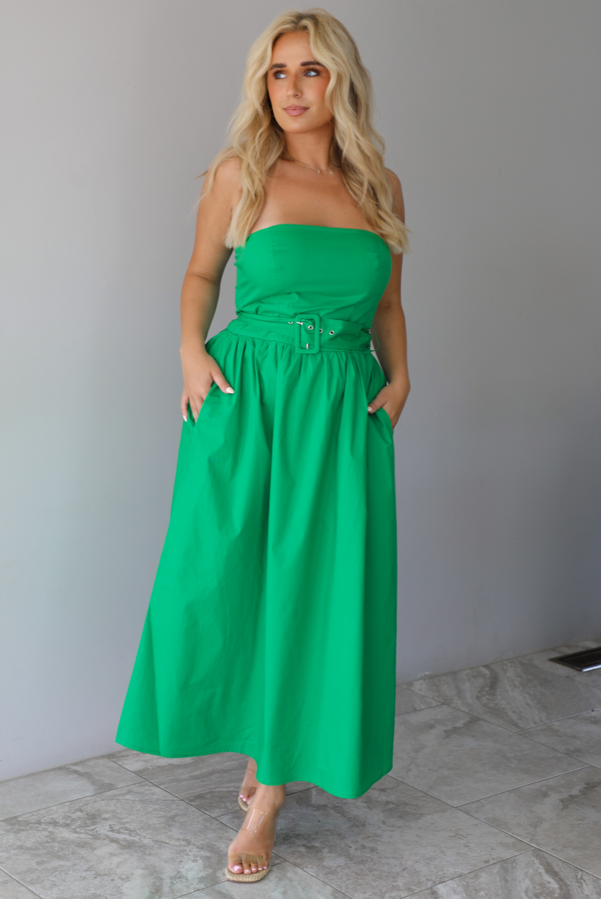Woman wearing a green strapless midi dress that has a fitted bodice, belted waistline detail, functional pockets on either side, and a midi length indoors against a white wall with white marble flooring. 