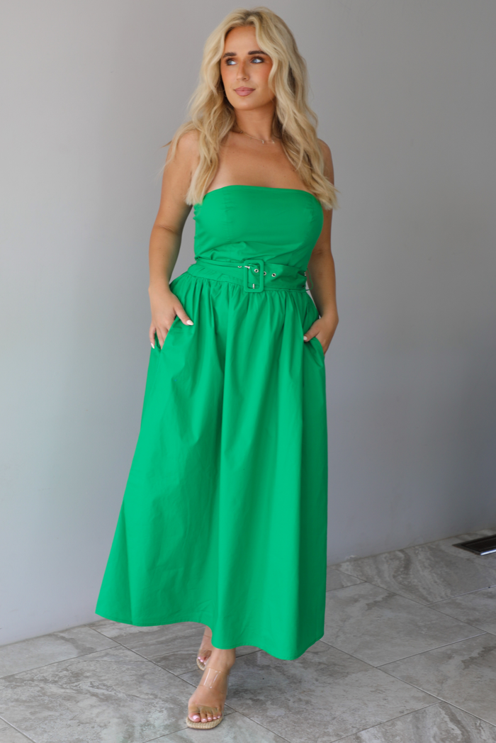 Woman wearing a green strapless midi dress that has a fitted bodice, belted waistline detail, functional pockets on either side, and a midi length indoors against a white wall with white marble flooring. 