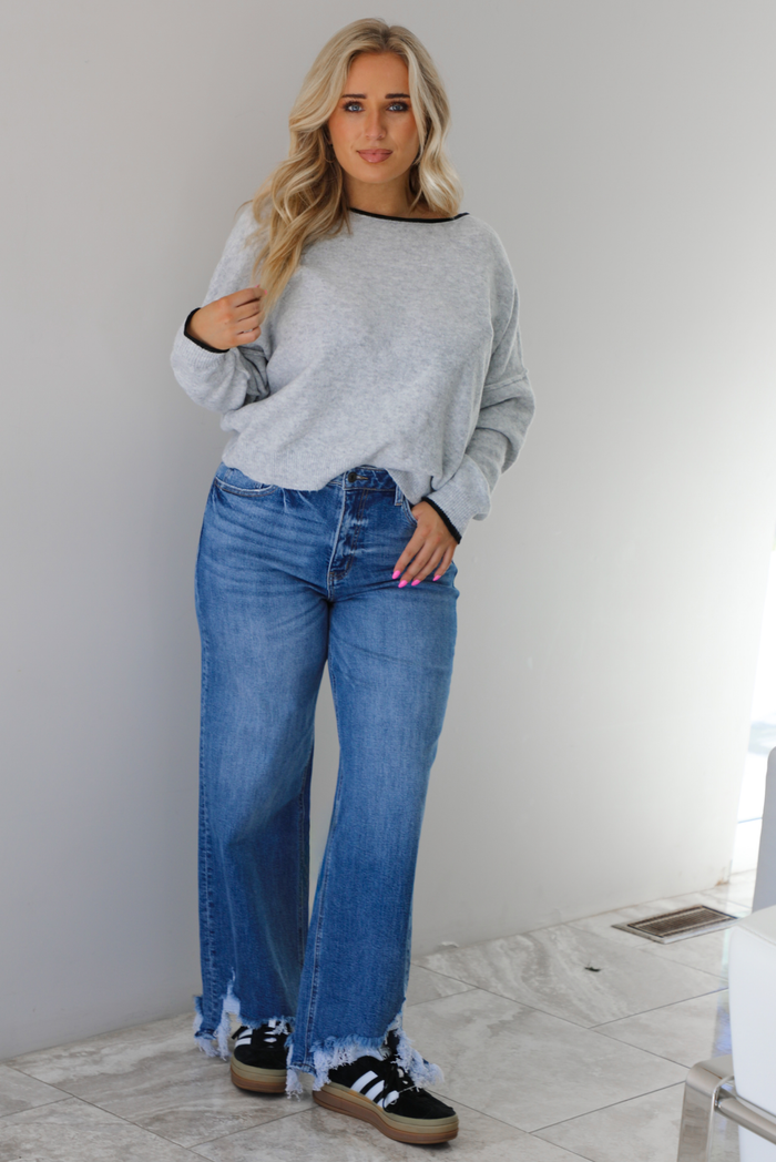 Woman wearing a grey relaxed fit sweater that has black trim on it, medium wash distressed denim jeans, and black sneakers. The photo is taken indoors against a white wall with white marble flooring. 