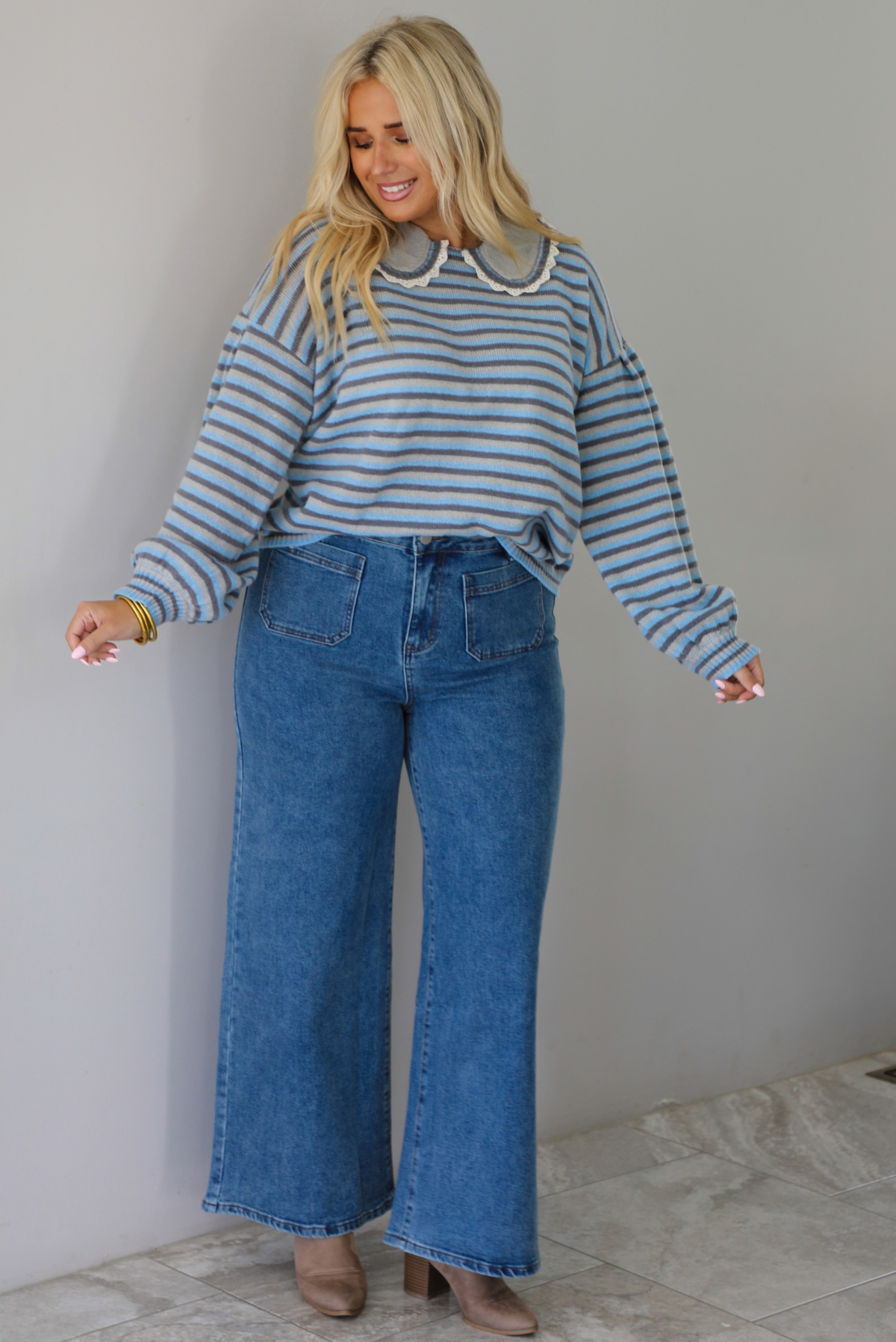 A woman modeling a light blue and navy striped long-sleeve sweater with a white ruffled collar, paired with wide-leg blue jeans and brown boots, standing indoors against a white wall with white marble flooring. 