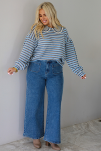 A woman modeling a light blue and navy striped long-sleeve sweater with a white ruffled collar, paired with wide-leg blue jeans and brown boots, standing indoors against a white wall with white marble flooring. 
