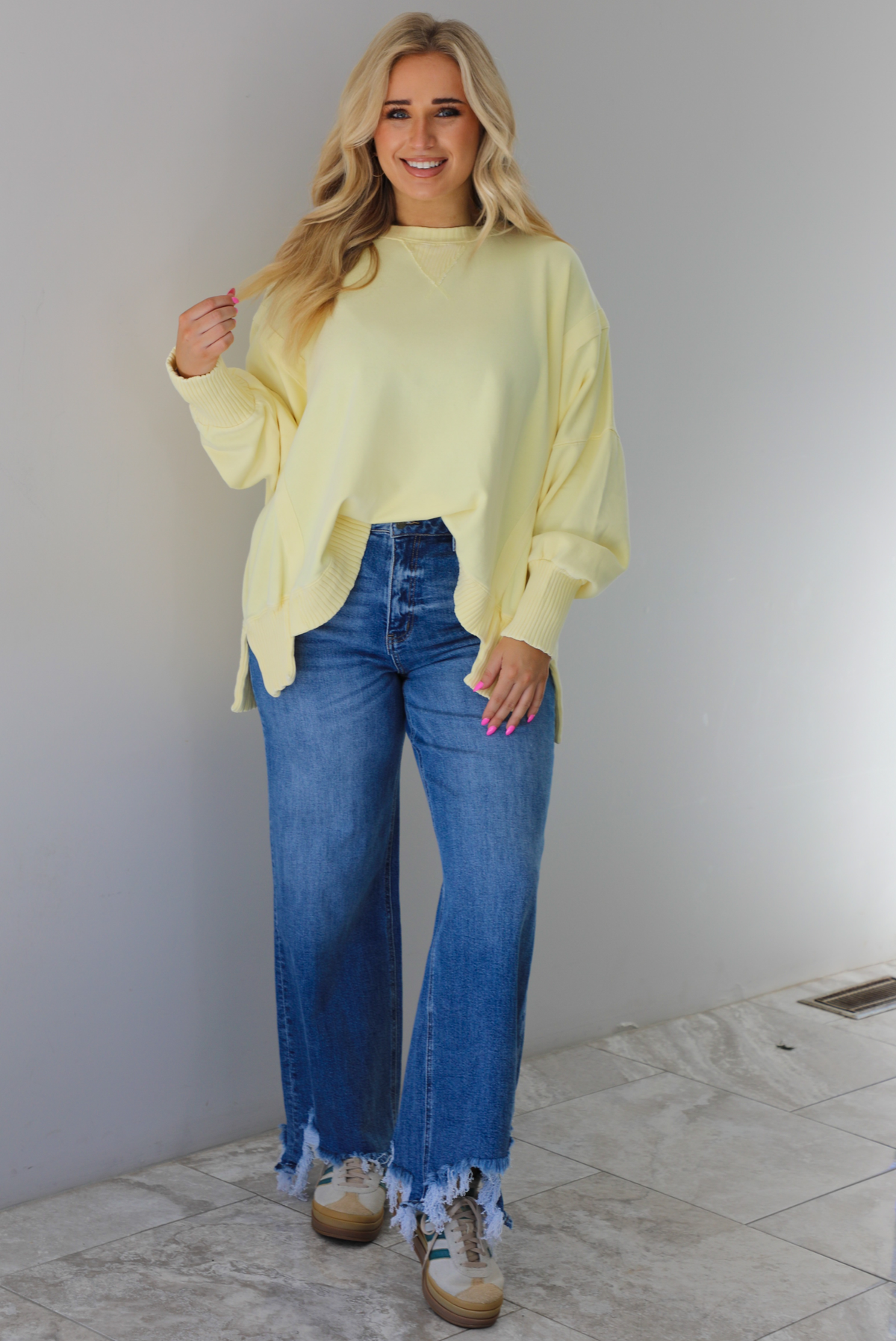 Woman wearing a pale yellow oversized sweater with dark wash denim and sneakers. The photo is taken indoors against a white wall with white marble flooring. 