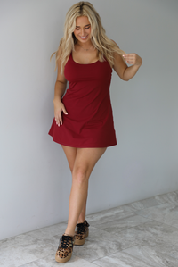 Woman wearing a maroon athleisure dress with thin straps and a relaxed fit indoors against a white wall with white marble flooring. 