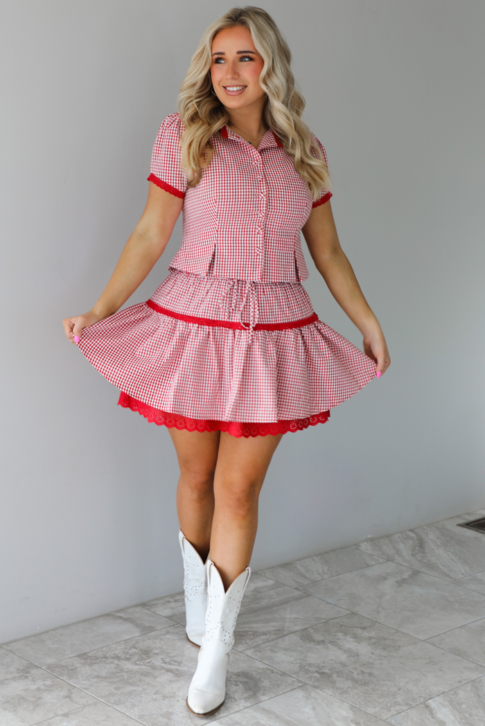 Woman wearing a red and white gingham patterned set that has a short sleeved button-up top that has pleated details at the hem of the top and a high waisted skirt with red lace detailing at the hem. The set is paired with white cowboy boots. The photo is taken indoors against a white wall with white marble flooring. 