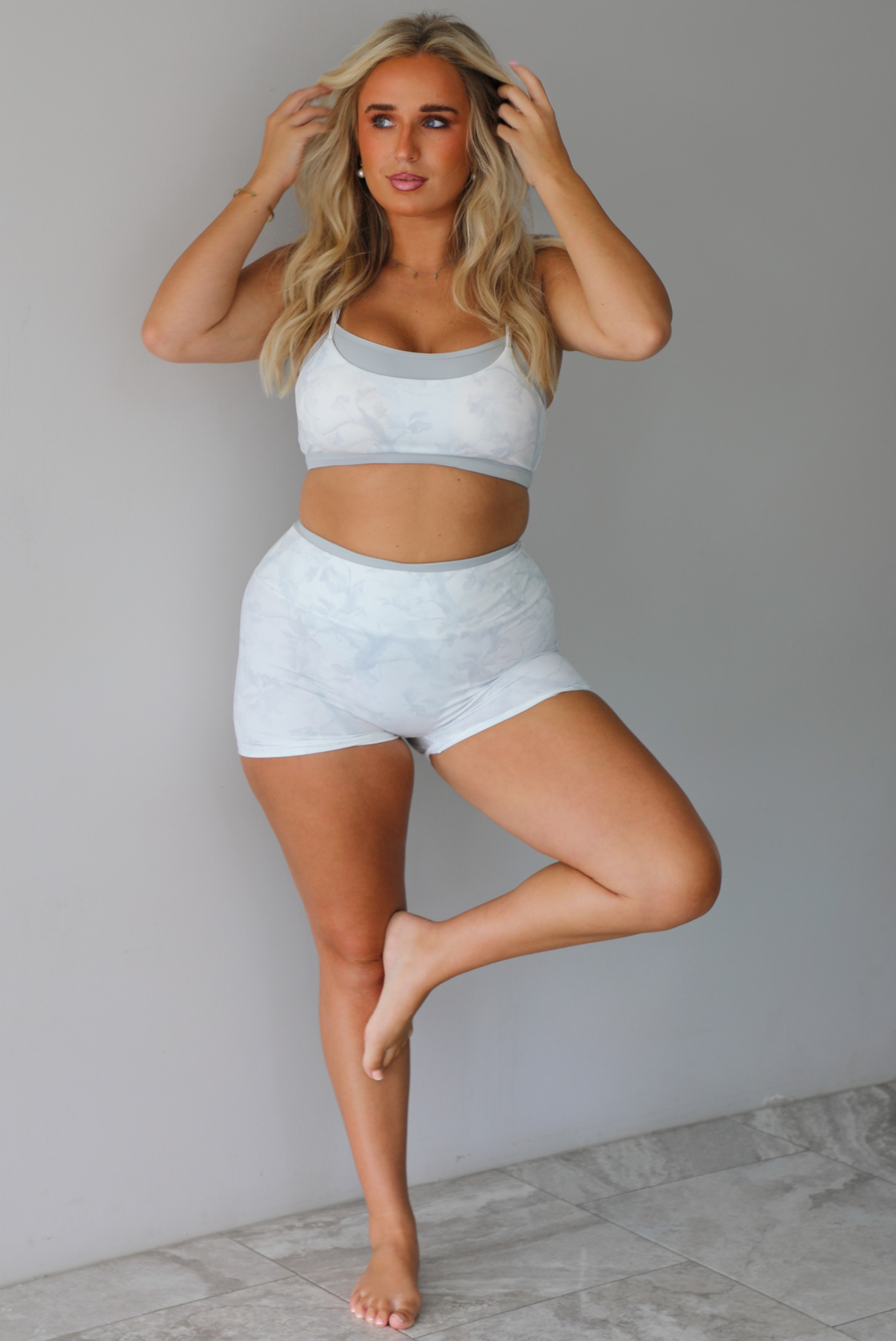 A woman wearing a matching grey and white camo activewear set consisting of a sports bra and high-waisted shorts, standing indoors against a white wall with white marble flooring. 