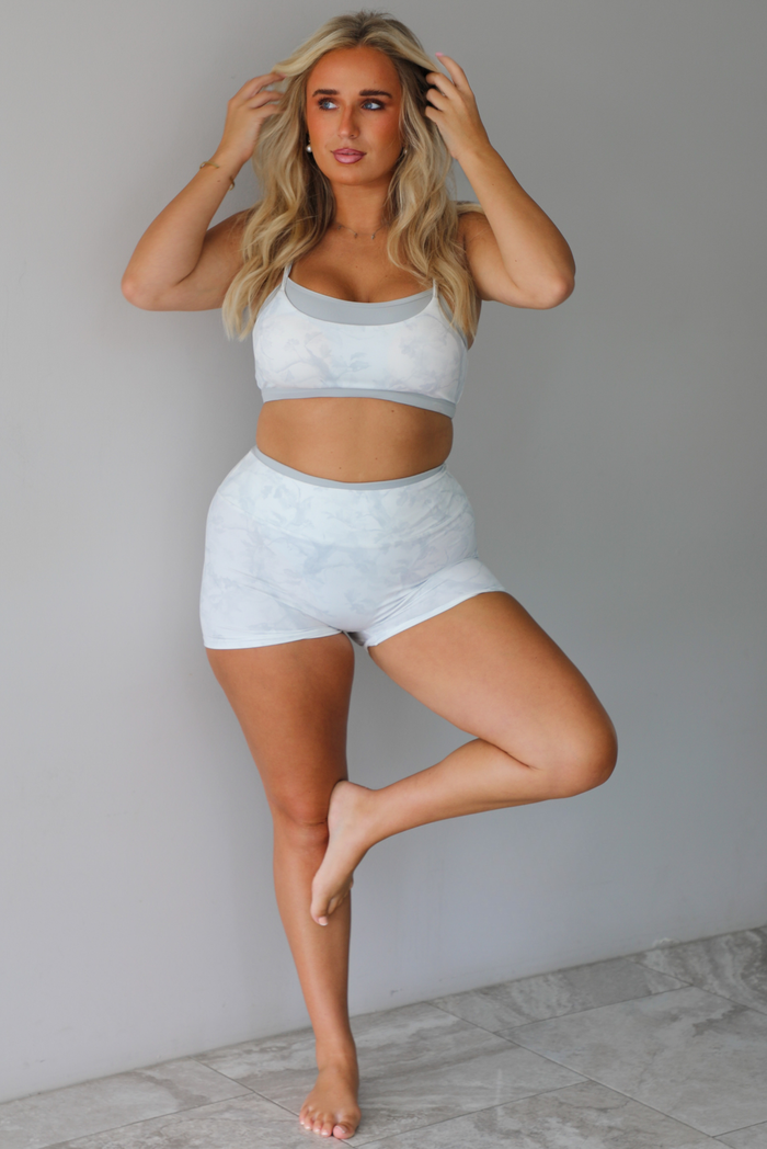 A woman wearing a matching grey and white camo activewear set consisting of a sports bra and high-waisted shorts, standing indoors against a white wall with white marble flooring. 