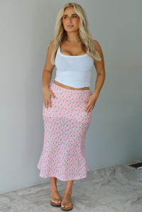 Woman wearing a white tank and pink midi length skirt that has a repeating floral pattern throughout indoors against a white wall with white marble flooring. 