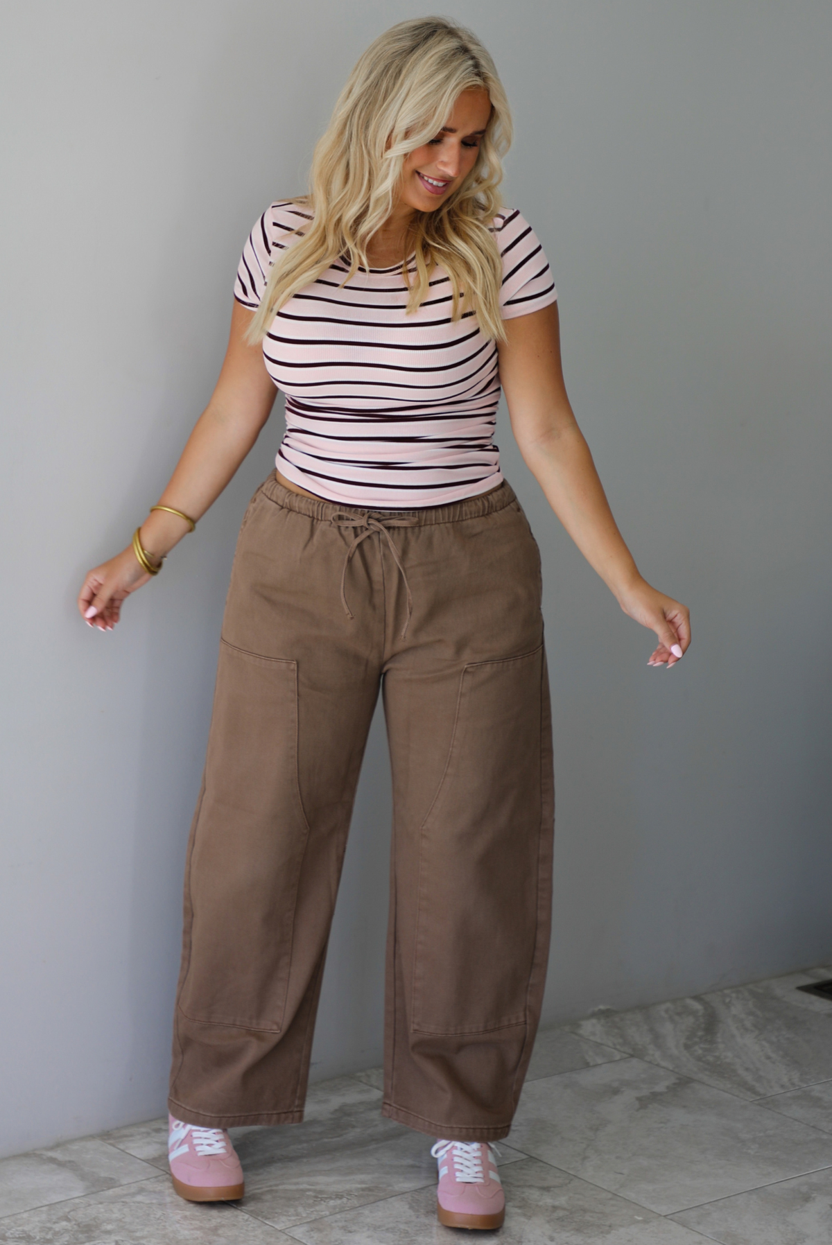 A woman wearing a pink baby tee that has a burgundy and white striped design throughout, paired with mocha barrel denim, standing indoors against a white wall with white marble flooring. 