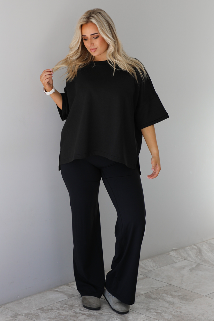 Woman wearing a black oversized box tee with longer short sleeves, a high-low design, and split hem details on either side. The tee is paired with black flared leggings. The photo is taken indoors against a white wall with white marble flooring. 