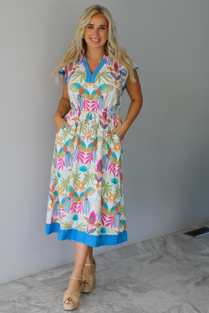 A woman wearing a short sleeved midi dress with a colorful tropical leaf print and light blue trim indoors against a white wall with white marble flooring. 