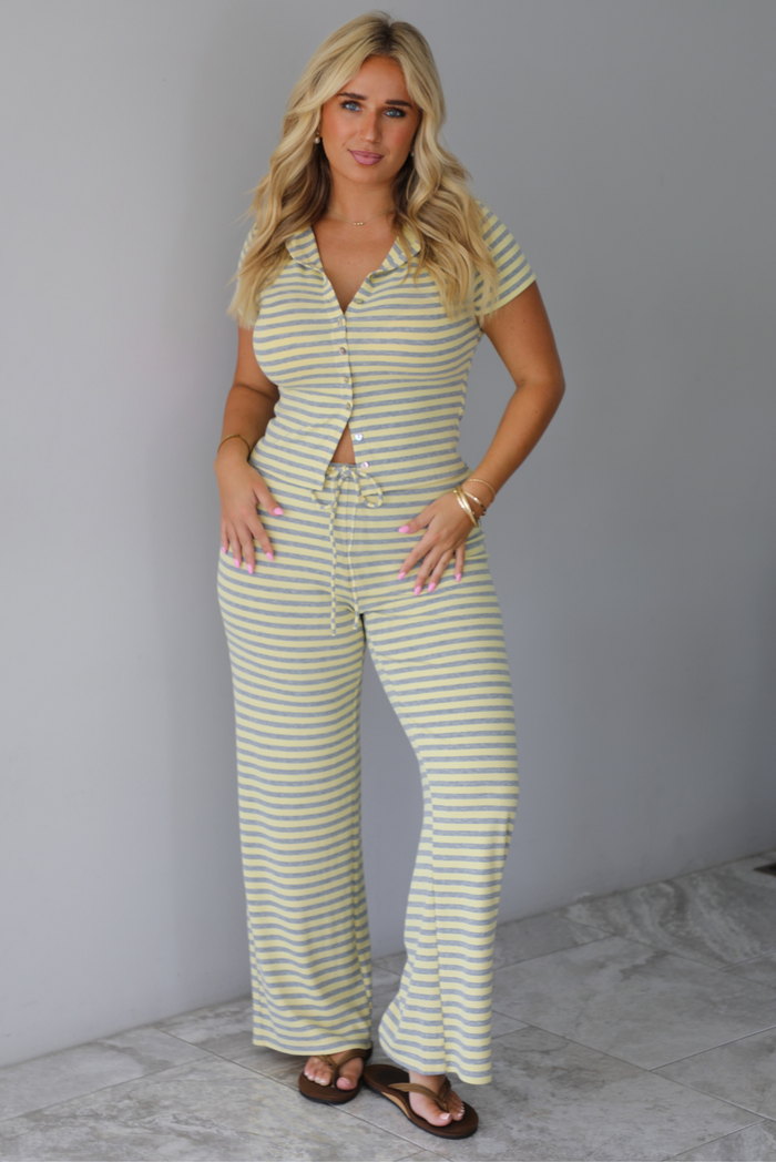 A woman wearing a two piece set consisting of a collared, button-down, short sleeve top and high waisted, drawstring, wide leg pants, standing indoors against a white wall with white marble flooring. 