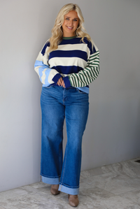 Woman wearing a navy, cream, blue, and green alternating striped design throughout, paired with dark denim jeans, standing indoors against a white wall with white marble flooring. 