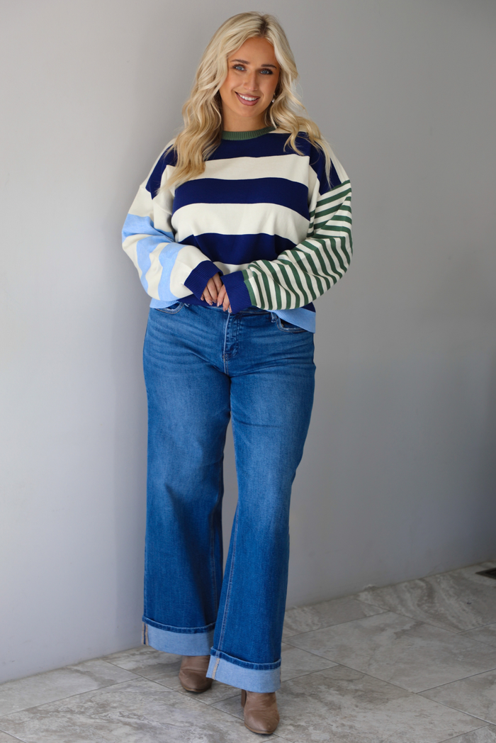 Woman wearing a navy, cream, blue, and green alternating striped design throughout, paired with dark denim jeans, standing indoors against a white wall with white marble flooring. 