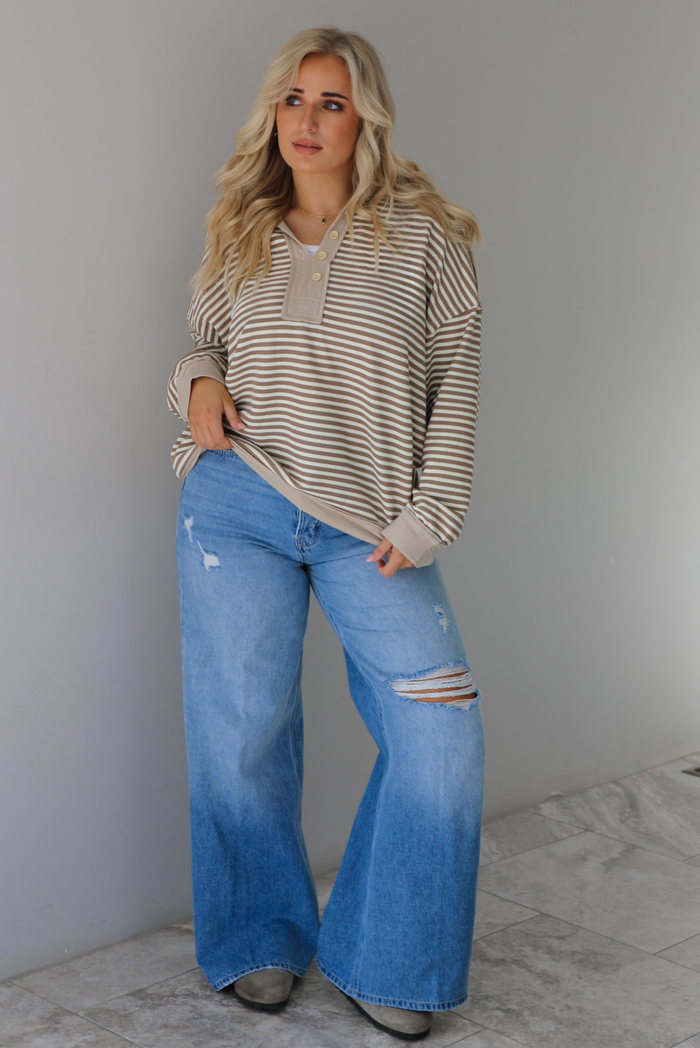 Woman wearing a mocha and cream striped pullover with a collared neckline, quarter button closures in the front, that has an oversized fit. The pullover is paired with a medium wash denim. The photo is taken indoors against a white wall with white marble flooring. 