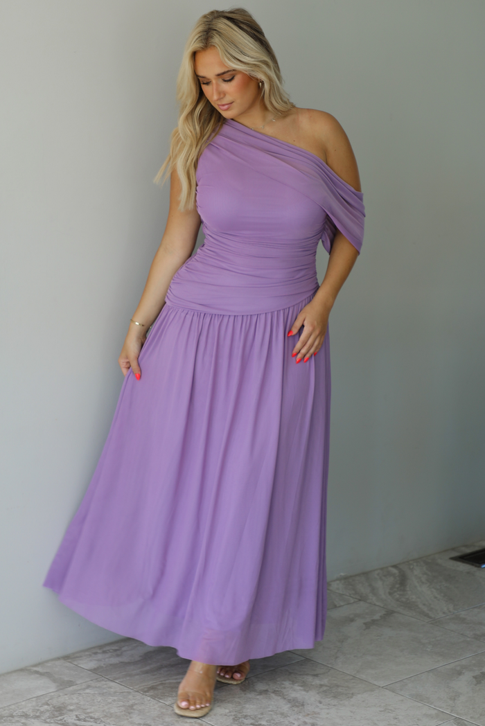 Woman wearing a lavender formal gown featuring a one strap that has extra mesh material that goes to the other arm and drapes down, has a fitted drop waist, and a long, maxi length skirt. The photo is taken indoors against a white wall with white marble flooring. 