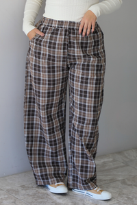 A woman shown from the waist down wearing a cream colored long sleeved top paired with brown plaid pants, standing indoors against a white wall with white marble flooring. 