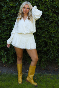 Woman model wearing an off-white romper with yellow boots outdoors in front of greenery. 
