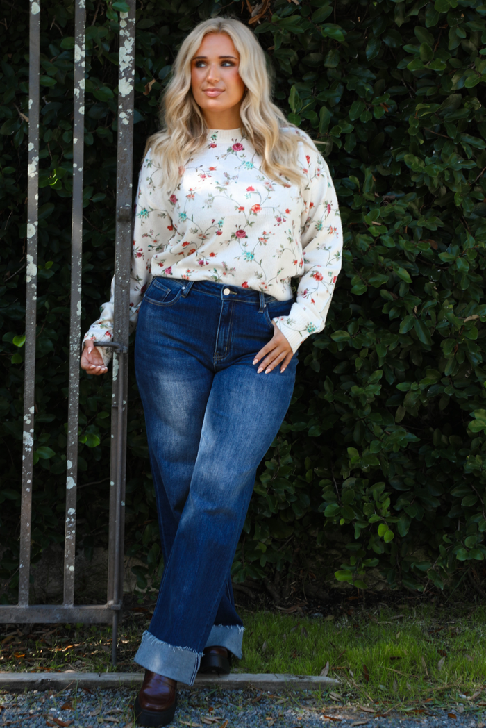 Woman model wearing an off-white sweater that has a multi-colored floral print pattern throughout with dark denim jeans. The photo is taken next to an iron gate outdoors with greenery in the back. 