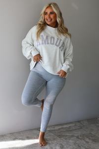 A woman posing in a light gray sweatshirt with 'AMOUR' printed in gray letters and light heather gray leggings, standing indoors against a white wall with white marble flooring. 