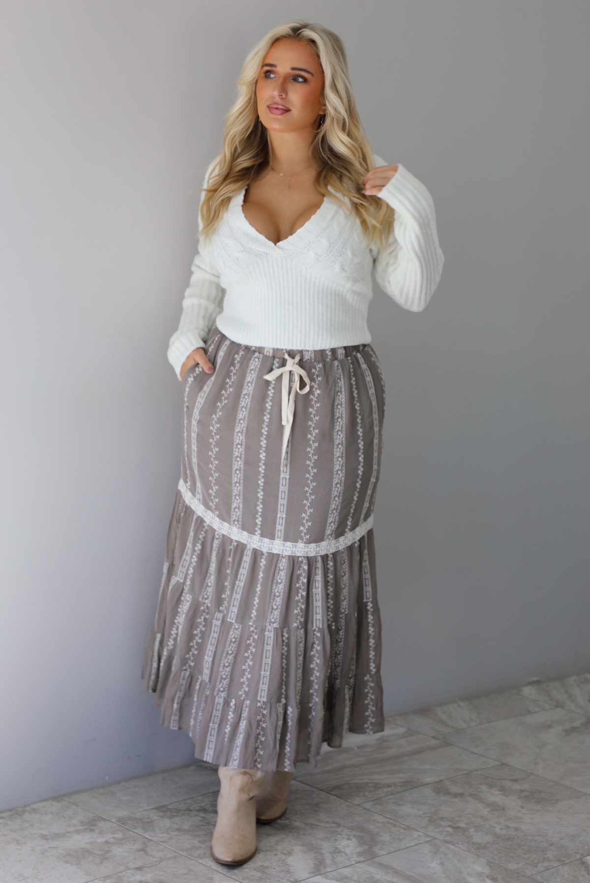 A woman wearing an ivory v-neck sweater paired with a taupe and white embroidered maxi skirt, standing indoors against a white wall with white marble flooring. 