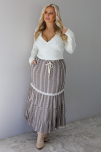 A woman wearing an ivory v-neck sweater paired with a taupe and white embroidered maxi skirt, standing indoors against a white wall with white marble flooring. 