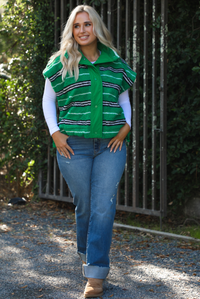 Woman model wearing a green, black, and white striped vest that has a sherpa texture to it, functional button closures in the front, and tie closures on either side, It is paired with a white long sleeved top, a medium wash pair of denim jeans, and tan ugg boots. The photo is taken outdoors with a metal gate and greenery in the back.