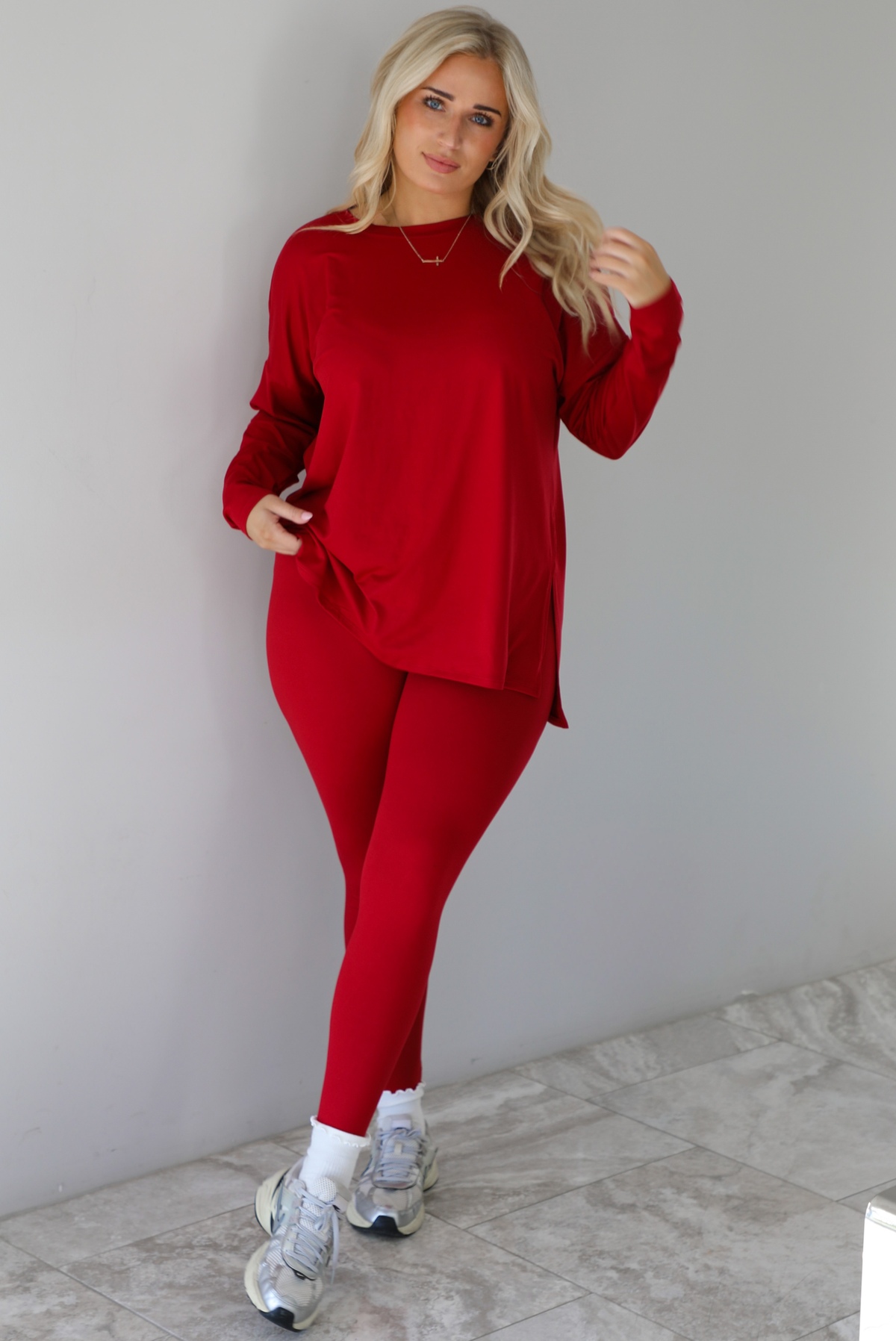 Woman wearing a red two piece set that features an oversized long sleeved top and fitted leggings. The set is paired with high ankle socks and silver sneakers. The photo is taken indoors against a white wall with white marble flooring.