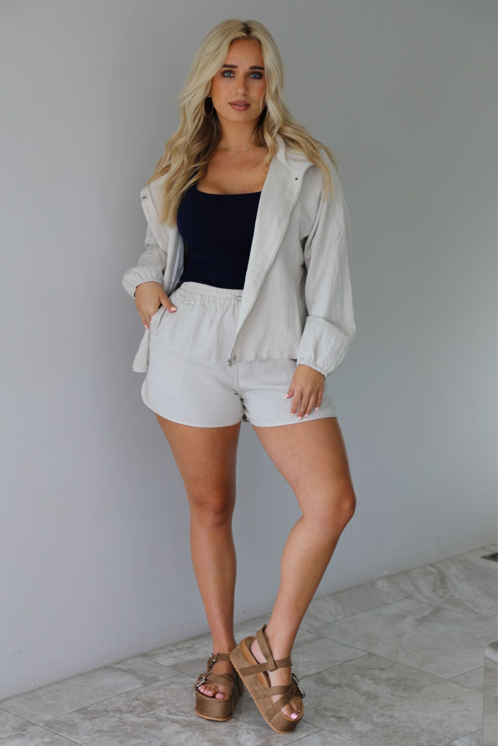 Woman wearing a stone colored two piece set featuring a zip up jacket and shorts with a navy tank tucked into the shorts indoors against a white wall with white marble flooring. 