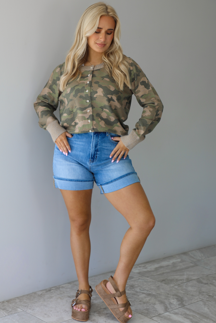 Woman wearing a camo cardigan with functional button closures in the front tucked into high waisted denim shorts. The photo is taken in front of a white wall with white marble flooring. 
