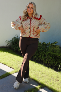 Woman model wearing a tan and red sherpa cardigan with a collared neckline and red trim with dark brown ahtleisure pants. The photo is taken outdoors with a white wall and greenery in the back. 