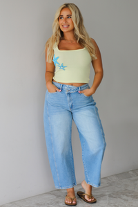 Woman wearing a yellow cropped tank with turquoise starfish studded details on the side with light wash barrel denim jeans, while standing indoors against a white wall with white marble flooring. 