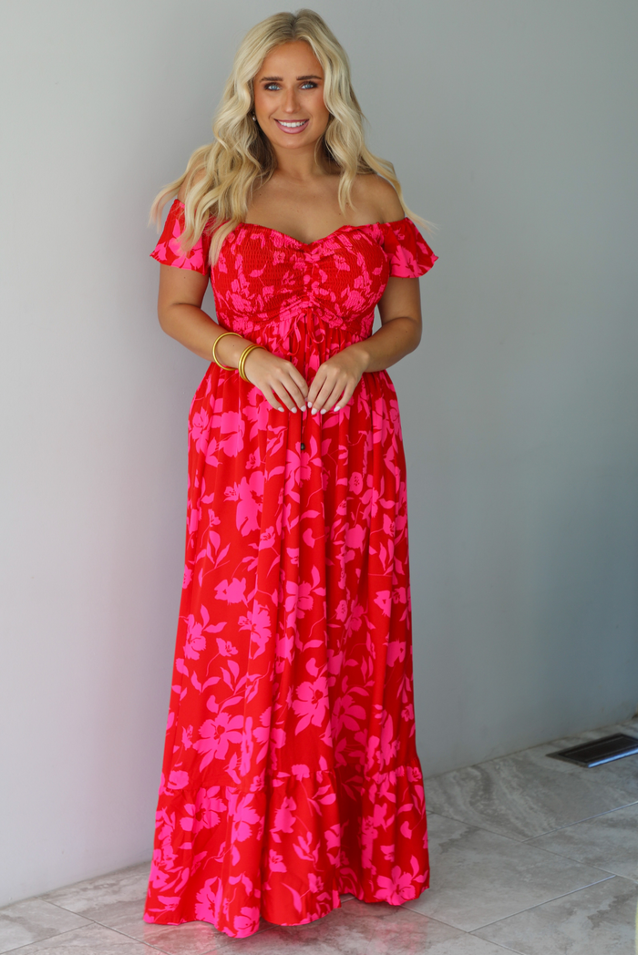 A woman modeling a bright red off-the-shoulder maxi dress with a vibrant pink floral pattern, standing indoors agains a white wall with white marble flooring. 
