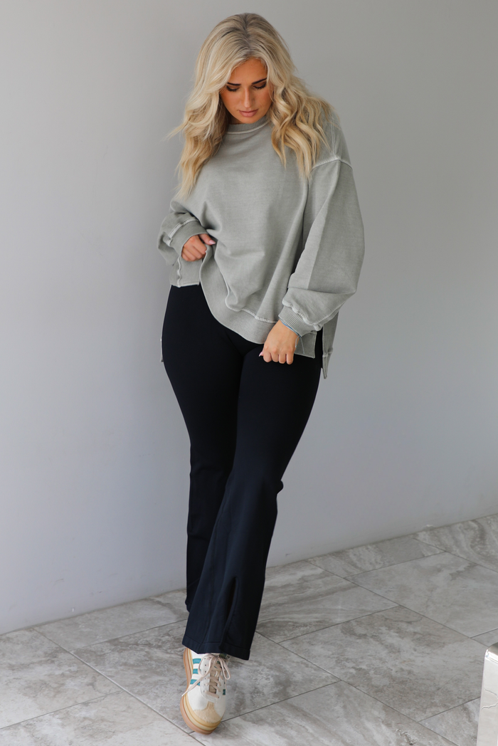 Woman wearing a tan oversized sweater with black leggings that have a slightly flared hem with split hem details in the front. The photo is taken indoors against a white wall with white marble flooring. 