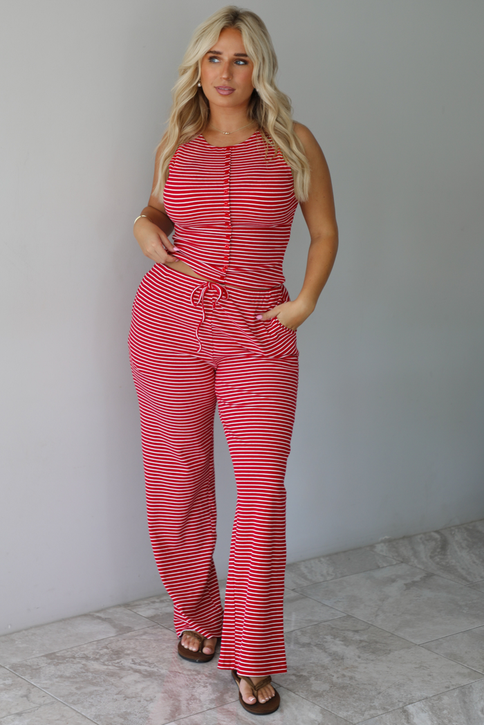 A woman modeling a red and white striped sleeveless set consisting of a tank top with non-functional button closures in the front and high waisted pants with a drawstring waistline and pockets on either side indoors against a white wall with white marble flooring. 