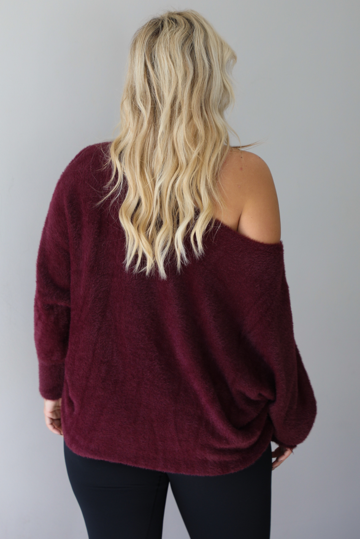 A woman wearing a maroon oversized long sleeved sweater, worn off the shoulder, paired with black leggings, indoors against a white wall.