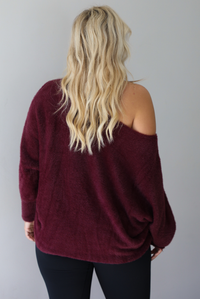 A woman wearing a maroon oversized long sleeved sweater, worn off the shoulder, paired with black leggings, indoors against a white wall.