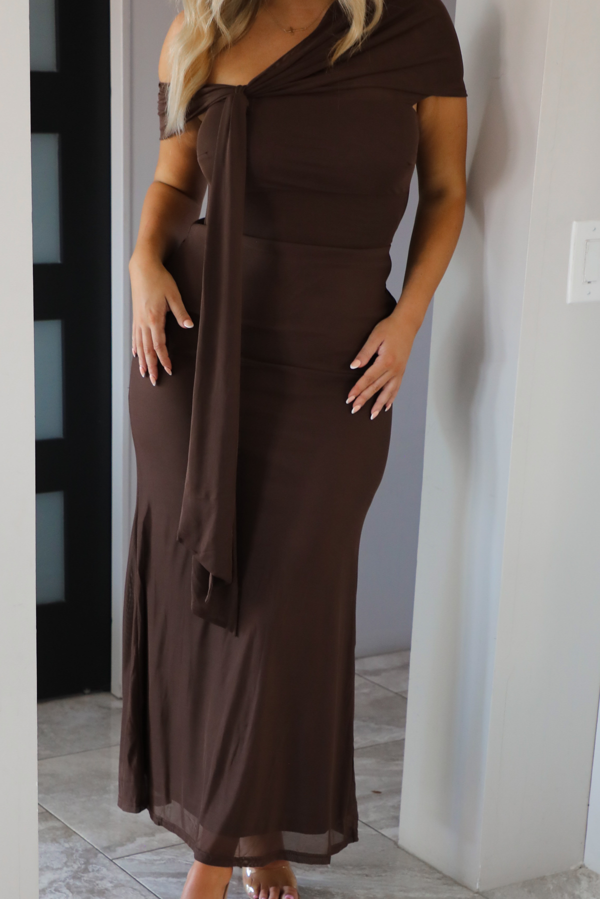 Woman model wearing a chocolate brown formal style dress that's fitted at the bodice, has asymmetrical sleeves, and a mermaid style skirt that's fitted at the hips and flared from the bottom of the high to the hem. The photo is taken inside a modern home in an archway. The photo showcases a close up of the dress.